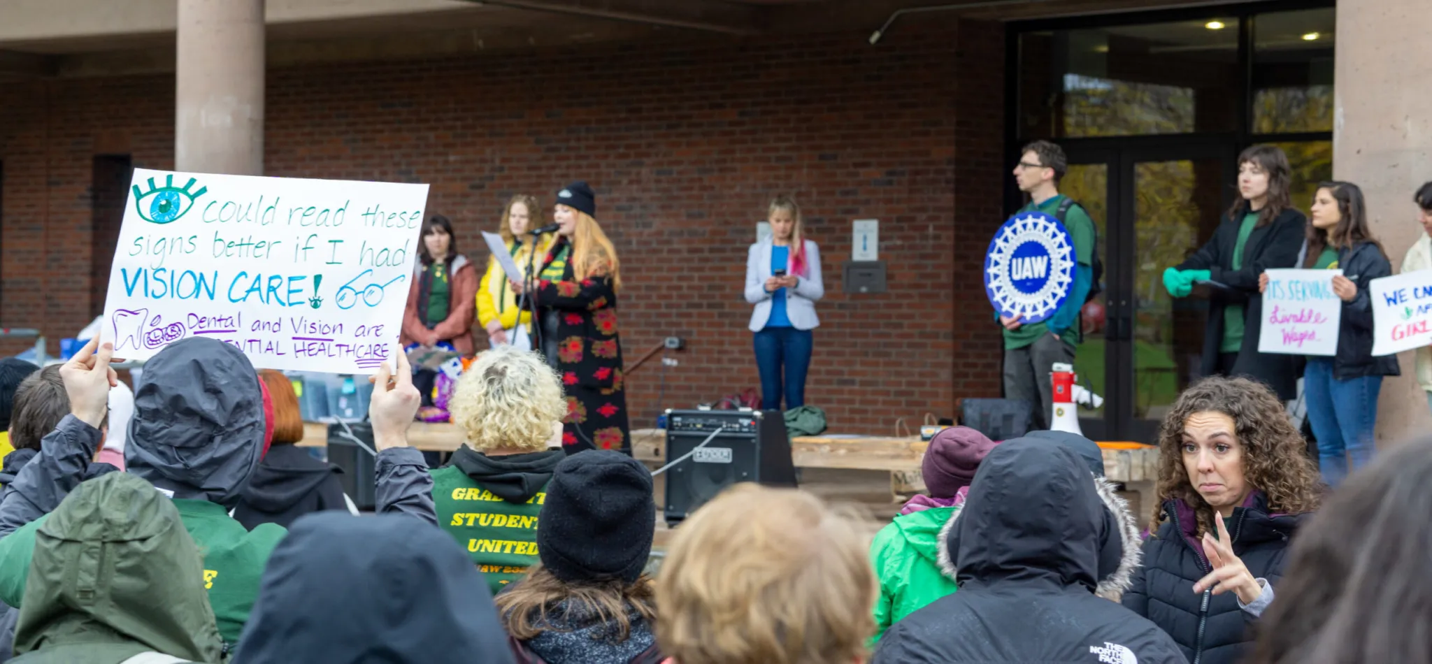 Graduate Students United rally at UVM, members holding signs reading 'Dental and Vision are essential healthcare' and 'I could read these signs better if I had VISION CARE'.